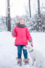 Fototapeta na wymiar Little girl in a bright jacket plays in the winter snowy forest with her dog