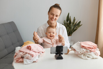 Indoor shot of smiling female blogger broadcasting livestream while sitting at table with her toddler baby and playing with her, using mobile phone for recording video or livestreaming, mom's blog.