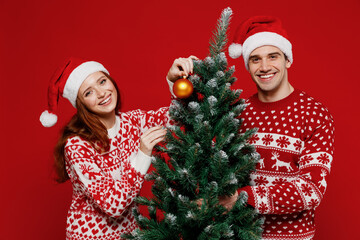 Young smiling happy couple friends two man woman 20s in sweater hat hold fir-tree decorating isolated on plain red background studio. Happy New Year 2022 celebration merry ho x-mas holiday concept