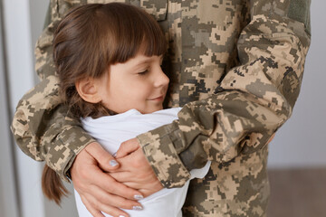 Indoor shot of cute charming girl hugging her mother who comes after war or from army, anonymous woman wearing military uniform embracing her child with love.