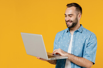 Young smiling happy freelancer copywriter satisfied caucasian man 20s wear blue shirt hold use work on laptop pc computer isolated on plain yellow background studio portrait. People lifestyle concept.