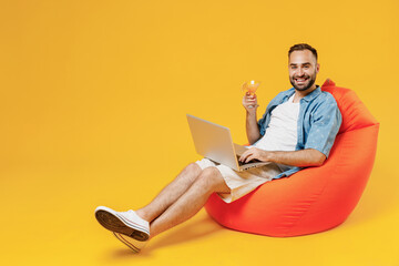 Full body young smiling cool happy man in blue shirt white t-shirt sit in bag chair hold use work on laptop pc computer alcohol cocktail martini isolated on plain yellow background studio portrait