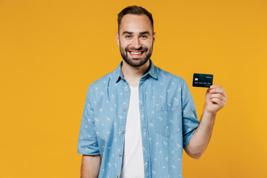 Young satisfied rich cool smiling happy caucasian man 20s wearing blue shirt white t-shirt hold in hand credit bank card isolated on plain yellow background studio portrait. People lifestyle concept.