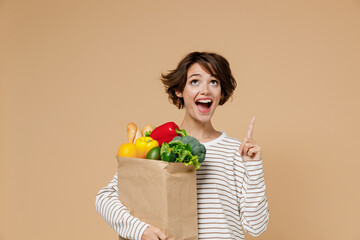 Young smiling happy vegetarian woman 20s in casual clothes hold paper bag with vegetables point finger overhead on workspace area copy soace mock up isolated on plain pastel beige background studio.