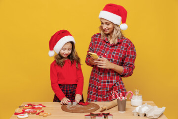 Two woman mother fun child baby girl in red Christmas hat isolated on plain yellow wall background studio. Mom little kid cook ginger cookie at kitchen table home. Happy New Year 2022 holiday concept