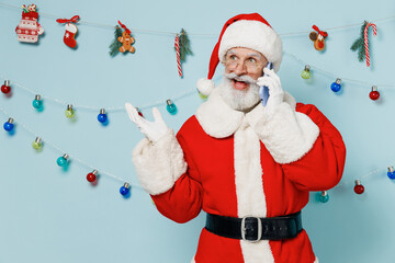 Old excited bearded Santa Claus man 50s wears Christmas hat red suit talk speak on mobile cell phone isolated on plain blue background studio. Happy New Year 2022 celebration merry ho x-mas concept.