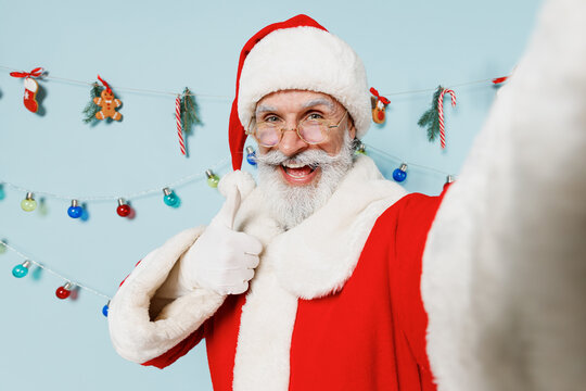 Close up old bearded Santa Claus man 50s in Christmas hat red suit do selfie shot pov on mobile phone show thumb up isolated on plain blue background studio Happy New Year 2022 merry ho x-mas concept.