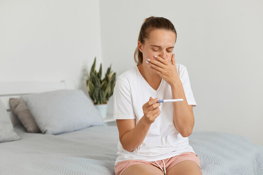 Pregnant attractive woman wearing casual attire sitting on sofa at home and holding pregnancy test with positive result, suffering from toxicosis and nausea.