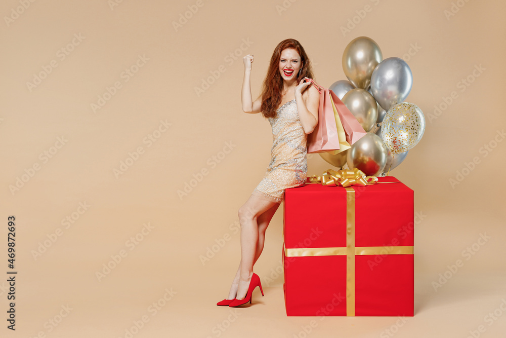 Wall mural Full size young woman in dress near balloons sit on big red gift present box hold package shopping bags do winner gesture isolated on plain pastel beige background. Celebrating birthday party concept.