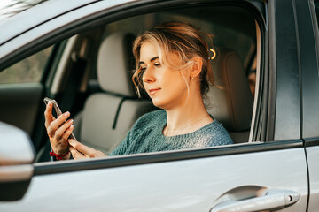 Naklejka premium Young beautiful woman sits behind wheel of car, looks at screen of mobile phone, typing sms massage uses smartphone, navigation system, map, assistance in finding out