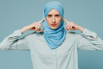 Young arabian asian muslim woman in abaya hijab cover ears with hands fingers do not want to listen scream isolated on plain blue background studio. People uae middle eastern islam religious concept.