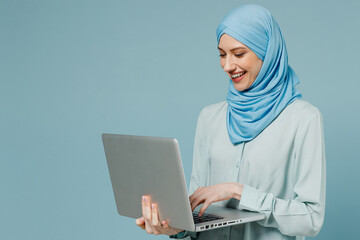 Young smiling happy arabian asian muslim woman in abaya hijab hold use work on laptop pc computer isolated on plain blue background studio portrait. People uae middle eastern islam religious concept. - obrazy, fototapety, plakaty