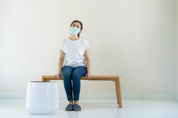 Cheerful Asian young woman sitting on the wooden bench beside the air purifier in the empty room with copy space. Woman wear a hygiene protective face mask, wellbeing and wellness concept.