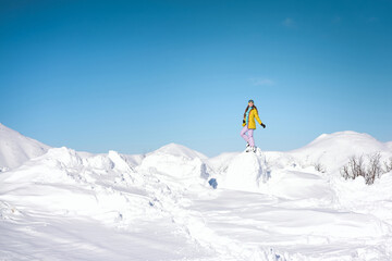 Cheerful girl snowboarder in yellow jacket in front of snowy mountains and blue sky