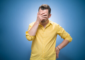 Young man covering face with hand. Shame over blue background, dresses in yellow shirt. Ashamed guy...