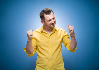 Winner man with clenched fist celebrating her success over blue background, dresses in yellow shirt. Cheerful male person wins lottery jackpots