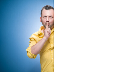 Hush. Man holding finger on lips, keep the secret concept over blue background, dresses in yellow shirt