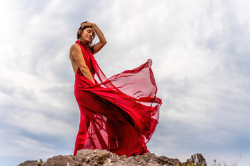 A woman in a red dress stands above a stormy sky, her dress fluttering, the fabric flying in the...
