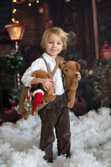 Cute fashion toddler boy, playing in the snow with teddy bear in front of a wooden cabin log