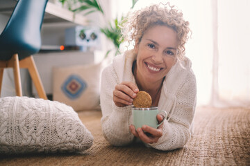 Happy adult young woman do breakfast at home laying on the carpet and eating cakes biscuits with...