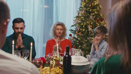 Loving family praying before Christmas dinner at home. Beautiful friends sitting at dining table set with delicious food during New Year party.