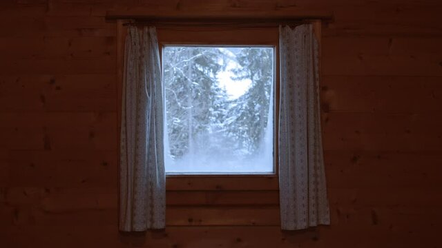 Blick durch ein kleines, zum Teil beschlagenes Fenster mit gemusterten Vorhängen einer Berghütte in den Alpen auf schneebedeckte Nadelbäume und friedlich herabfallende Schneeflocken. 