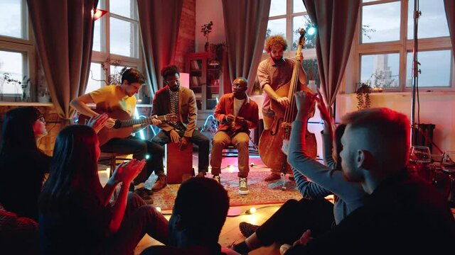 Slow motion arc shot of multiethnic music band giving live concert in studio with neon light while people clapping hands to them