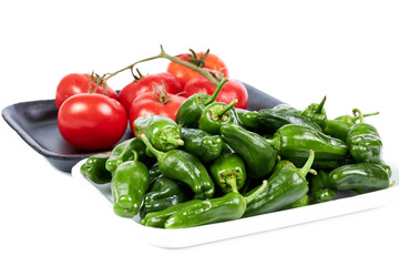 Red vine tomatoes on a black tray and green peppers on a white tray on a white background