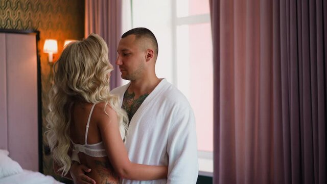 Man in white bathrobe hugs attractive blonde lady in lacy bra standing by large bed in comfortable hotel room at honeymoon slow motion