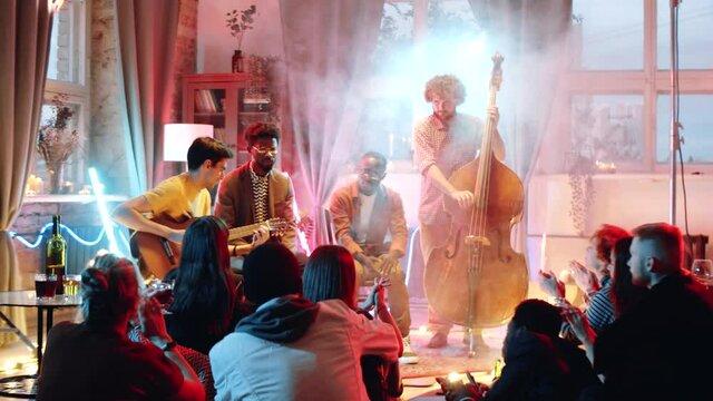 Young diverse musicians playing guitar and double bass during small atmospheric concert in studio with smoke and neon light