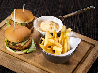 two small hamburgers with pickles served on cutting board with french fries and sauce thousand island on dark background
