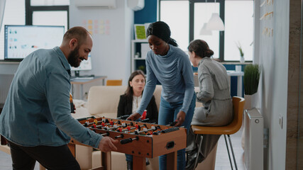 Man playing soccer game with woman at foosball table, meeting for drinks and entertainment at office after work. Multi ethnic group of colleagues enjoying beer and pizza after hours