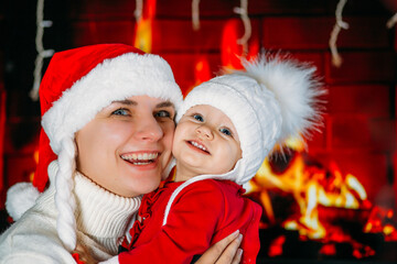 Mom in a New Year's hat holds a daughter in a red outfit in her arms against the background of a fireplace