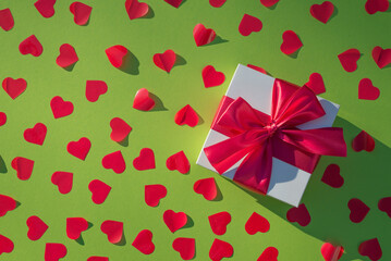 Fototapeta na wymiar White gift box with red ribbon on a green background. Valentines day backdrop with red confetti in the form of hearts. Flat lay style with minimalistic design. Template for banner or party invitation