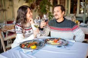 Happy young multi-ethnic couple having Christmas dinner in a rustic restaurant, talking to each other and drinking wine