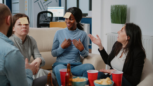 African american woman trying to guess game with sticky notes after work. Colleagues playing guessing game to do fun activity while celebrating party with beer and pizza at office.