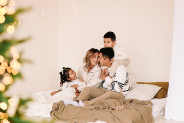 A mixed race Asian family with two children in cozy sweaters is relaxing together, resting and having fun on a bed in a decorated bedroom with Christmas tree at home in the New Year. Selective focus