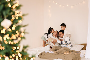 A mixed race Asian family with two children in cozy sweaters is relaxing together, resting and having fun on a bed in a decorated bedroom with Christmas tree at home in the New Year. Selective focus