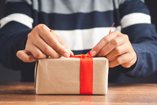 Man in sweater wrapping a gift on wooden table. A parcel wrapped in brown paper and tied with a red ribbon. Close-up photo. Concept of Christmas, New Year, Anniversary, Happy birthday, etc