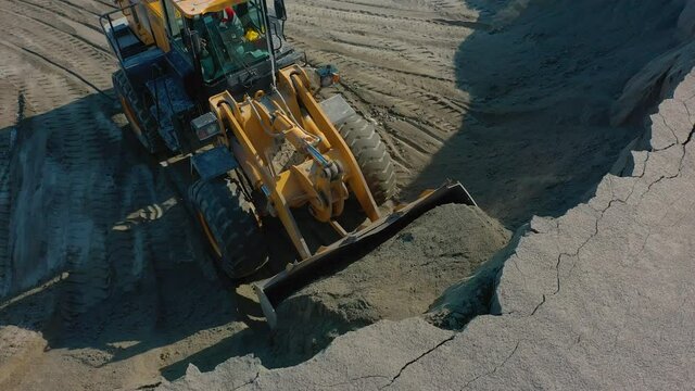 Wheel loader scoops up crushed stone with bucket from pile of sand to then load into dump truck. Heavy machinery working in quarry, aerial drone shot, close up shot. Excavator working