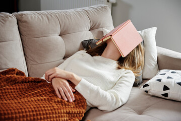 Tired woman sleeps on couch with book covered her face. Leisure day concept. Caucasian woman relaxing on sofa