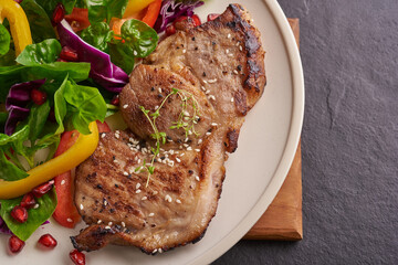Grilled steak with fresh vegetable, sweet pepper, tomatoes, red onion, pink pepper and spices. Home made tasty food. Concept for tasty and healthy meal. Black stone background. Pork steak with salad