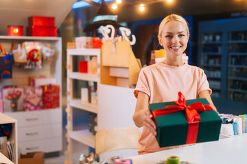 Cheerful attractive young woman offering take Christmas gift box tied red ribbon and decorated beautiful bow standing at counter of holiday store, bright blurred background of festive packages.