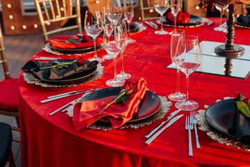 Guest table setting for banquet in black, red and gold style. Elegant and luxury dinner arrangement: decor, tablecloth, plates with napkins and fresh roses, glasses, cutlery. Themed party celebration