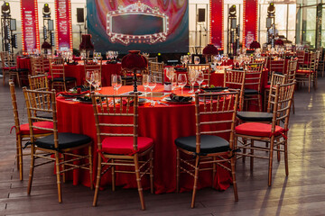 Guest's tables and chairs arrangement for banquet in black, red and gold style. Elegant event hall: decor, tablecloth, plates, napkins, roses, glasses. Themed party celebration on the roof, outdoor.