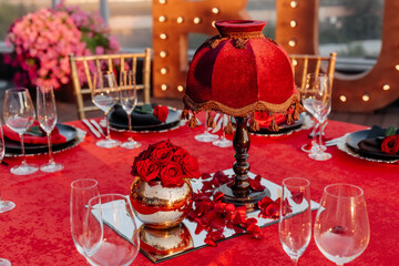 Guests table setting for banquet in black, red and gold style. Elegant dinner: decor, tablecloth, plates with napkins and fresh roses, glasses, cutlery. Themed party celebration on the roof, outdoor.