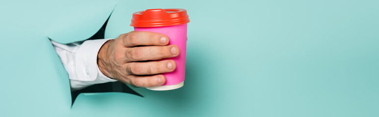 cropped view of man holding coffee to go through hole in paper wall on blue, banner