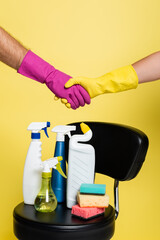 cropped view of cleaners shaking hands near reaching bottles with detergent and clean sponges on chair isolated on yellow