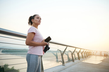 Determined sportswoman in pink T-shirt and gray sports leggings with smartphone in her hands and looking into the distance, standing on the treadmill of a city bridge, ready for a morning run at dawn