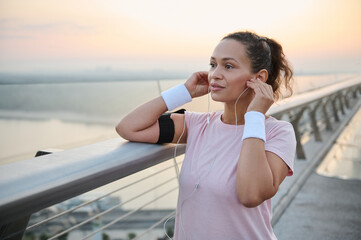 Beautiful sporty African American woman in sportswear putting on earphones, listening to music, getting ready for morning jog along the city bridge. Sportswoman at workout outdoor at daybreak